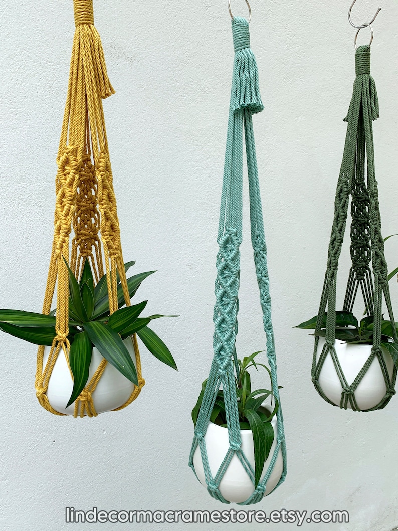 May include: Three macrame plant hangers in mustard yellow, seafoam green, and olive green, each holding a white ceramic pot with green plants. The hangers are suspended against a white wall. The Etsy shop name "lindecormacramestore.etsy.com" is visible at the bottom.