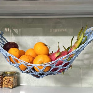 May include: A light blue macrame fruit hammock holding oranges, dragon fruit, and a dark purple fruit. The hammock is suspended from a metal shelf. Jars of nuts and seeds are on the counter below.