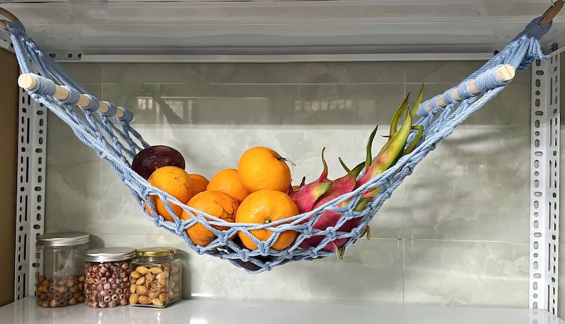 May include: A light blue macrame fruit hammock holding oranges, dragon fruit, and a dark purple fruit. The hammock is suspended from a metal shelf. Jars of nuts and seeds are on the counter below.