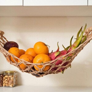 May include: A woven fruit hammock holding oranges, dragon fruit, and a plum. The hammock is suspended from a white cabinet. Jars of nuts and seeds are visible below.