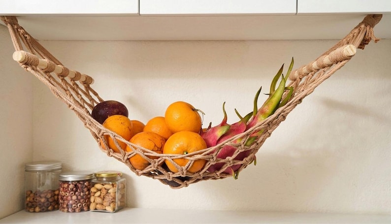 May include: A woven fruit hammock holding oranges, dragon fruit, and a plum. The hammock is suspended from a white cabinet. Jars of nuts and seeds are visible below.