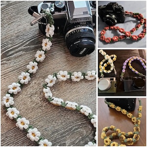 May include: A camera strap featuring a repeating pattern of crocheted white daisy flowers with yellow centers, connected by a green cord. The strap includes metal swivel clips at each end for attachment to a camera body, showcasing a floral design.