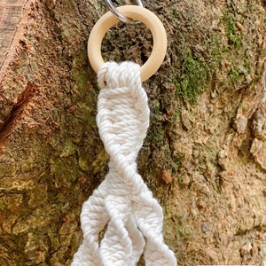 May include: A white macrame plant hanger with a wooden ring at the top. The hanger is hanging from a metal hook on a tree trunk.