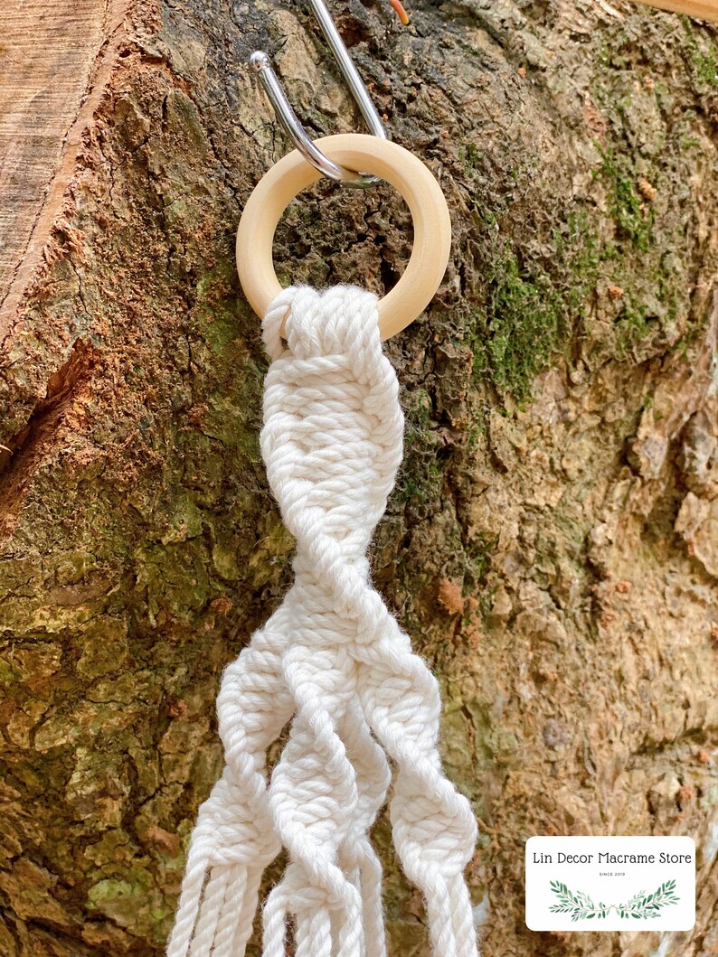 May include: A white macrame plant hanger with a wooden ring at the top. The hanger is hanging from a metal hook on a tree trunk.
