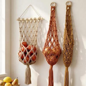 Puede incluir: Tres fruteros de macramé en beige, naranja y amarillo mostaza cuelgan de una pared blanca. El soporte beige, suspendido de un listón de madera, contiene tomates rojos. Los soportes naranja y mostaza, con aros de madera, contienen cebollas.