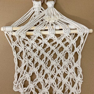 May include: Cream-coloured macrame wall hanging with a diamond-patterned net design. The top features two wooden rings and a wooden dowel. The macrame is made of thick, woven cotton rope. The Etsy shop name is visible at the bottom.