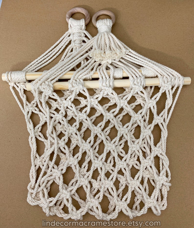 May include: Cream-coloured macrame wall hanging with a diamond-patterned net design. The top features two wooden rings and a wooden dowel. The macrame is made of thick, woven cotton rope. The Etsy shop name is visible at the bottom.