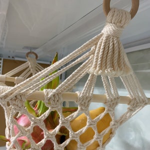 May include: Cream-coloured macrame fruit hammock with wooden dowels and a wooden ring for hanging. The hammock is filled with fresh fruit, including dragon fruit and mangoes. The text "lindecormacramestore.etsy.com" is visible.