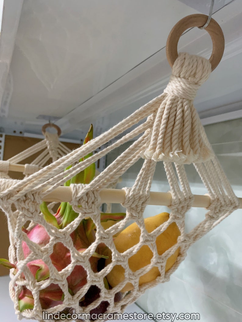 May include: Cream-coloured macrame fruit hammock with wooden dowels and a wooden ring for hanging. The hammock is filled with fresh fruit, including dragon fruit and mangoes. The text "lindecormacramestore.etsy.com" is visible.