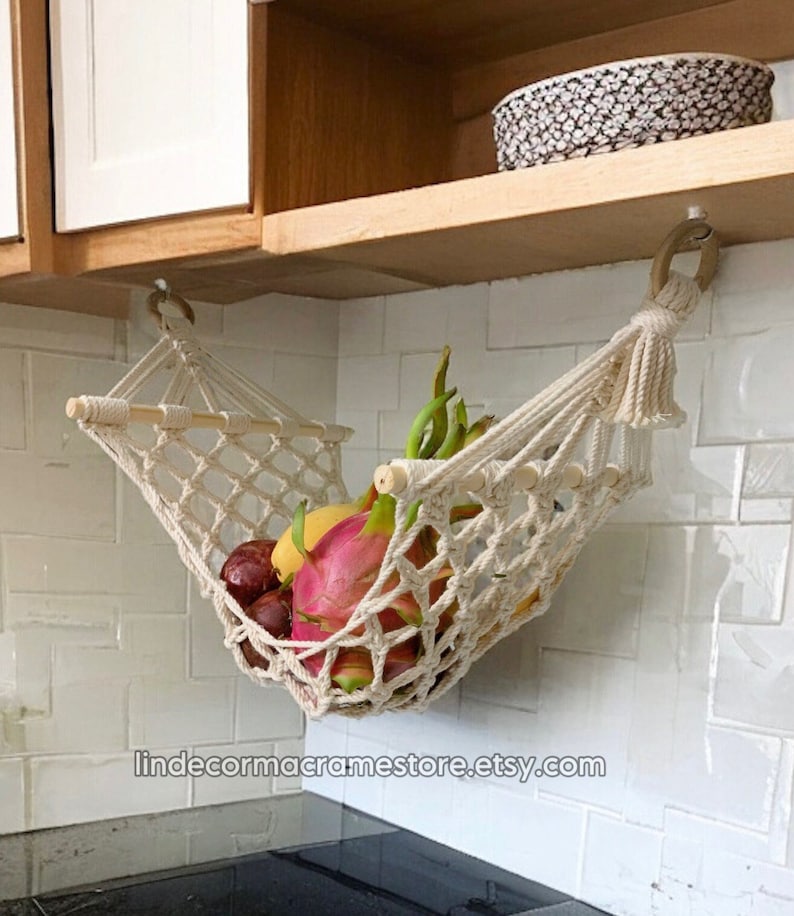 May include: A cream-coloured macrame fruit hammock, filled with a variety of colourful fruit, including dragon fruit and mangoes. The hammock is suspended from a wooden rod against a white tiled wall. The Etsy shop URL is visible.