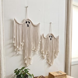May include: Two off-white macrame wall hangings shaped like ghosts, each featuring black felt eyes and a mouth. The ghost figures are constructed from long, vertical cotton cords with knotted ends, suspended from a single cord loop at the top.