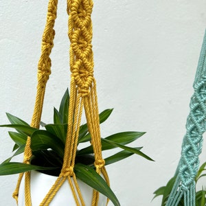 May include: Mustard yellow macrame plant hanger with a white ceramic pot holding a green plant. The macrame design features intricate knotting patterns. The Etsy shop URL is visible at the bottom of the image.