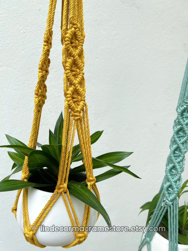 May include: Mustard yellow macrame plant hanger with a white ceramic pot holding a green plant. The macrame design features intricate knotting patterns. The Etsy shop URL is visible at the bottom of the image.