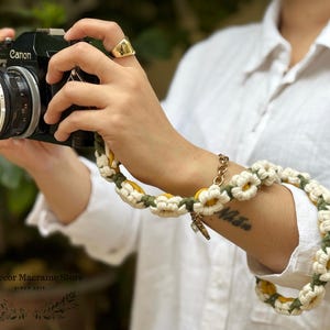 May include: A black Canon camera attached to a handmade crochet camera strap featuring a repeating pattern of white daisy flowers with yellow centers, connected by dark green yarn links. The strap is wrapped around a wrist for support.