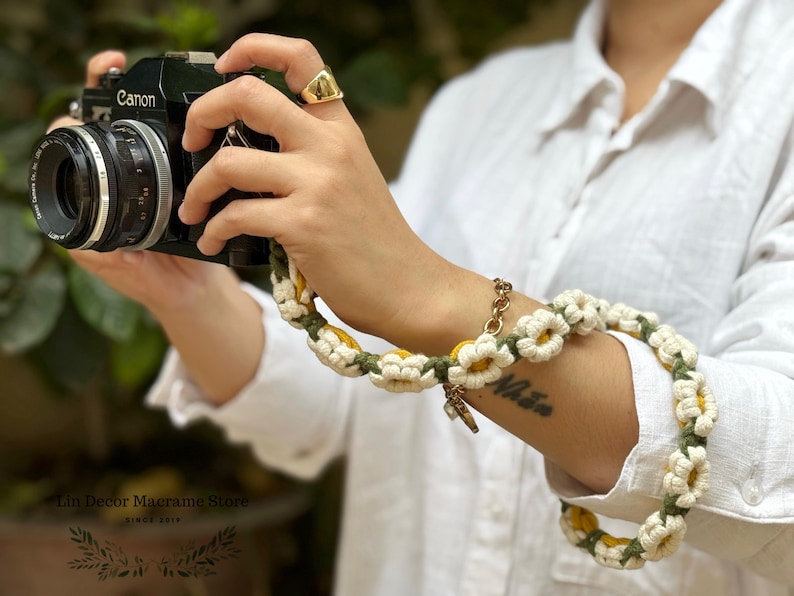 May include: A black Canon camera attached to a handmade crochet camera strap featuring a repeating pattern of white daisy flowers with yellow centers, connected by dark green yarn links. The strap is wrapped around a wrist for support.