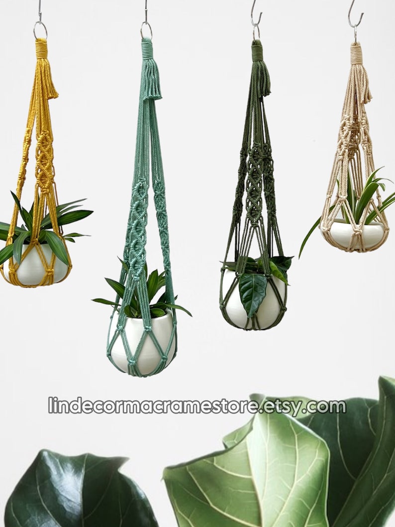 May include: Four macrame plant hangers in yellow, teal, olive green, and beige, each holding a white ceramic pot with a green plant. The hangers are suspended from metal hooks against a white background. The Etsy shop name is visible at the bottom.