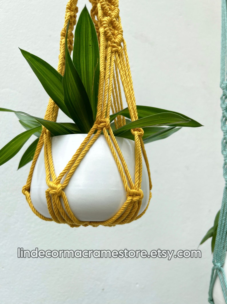 May include: A yellow macrame plant hanger holds a white ceramic pot with green leafy plants. The macrame is made of thick, twisted rope. The Etsy shop name is visible at the bottom of the image.