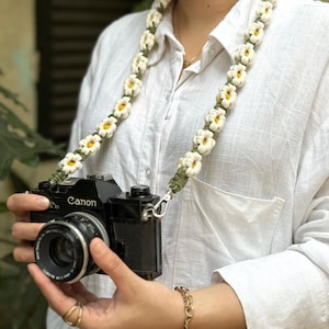 May include: A camera strap featuring a series of crocheted white daisy flowers with yellow centers connected by a dark green cord. The strap is attached to a black Canon camera body with a lens, showcasing a floral design and metal hardware.