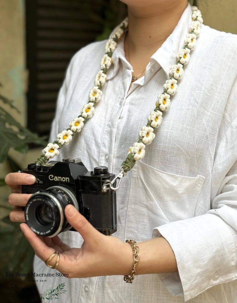 May include: A camera strap featuring a series of crocheted white daisy flowers with yellow centers connected by a dark green cord. The strap is attached to a black Canon camera body with a lens, showcasing a floral design and metal hardware.