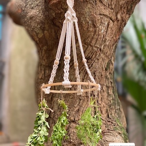 May include: A white macrame hanging planter with a wooden ring and wooden beads. The planter is hanging from a tree branch and has three bundles of dried herbs hanging from it. The text "Lin Decor Macrame Store" is visible in the bottom right corner of the image.