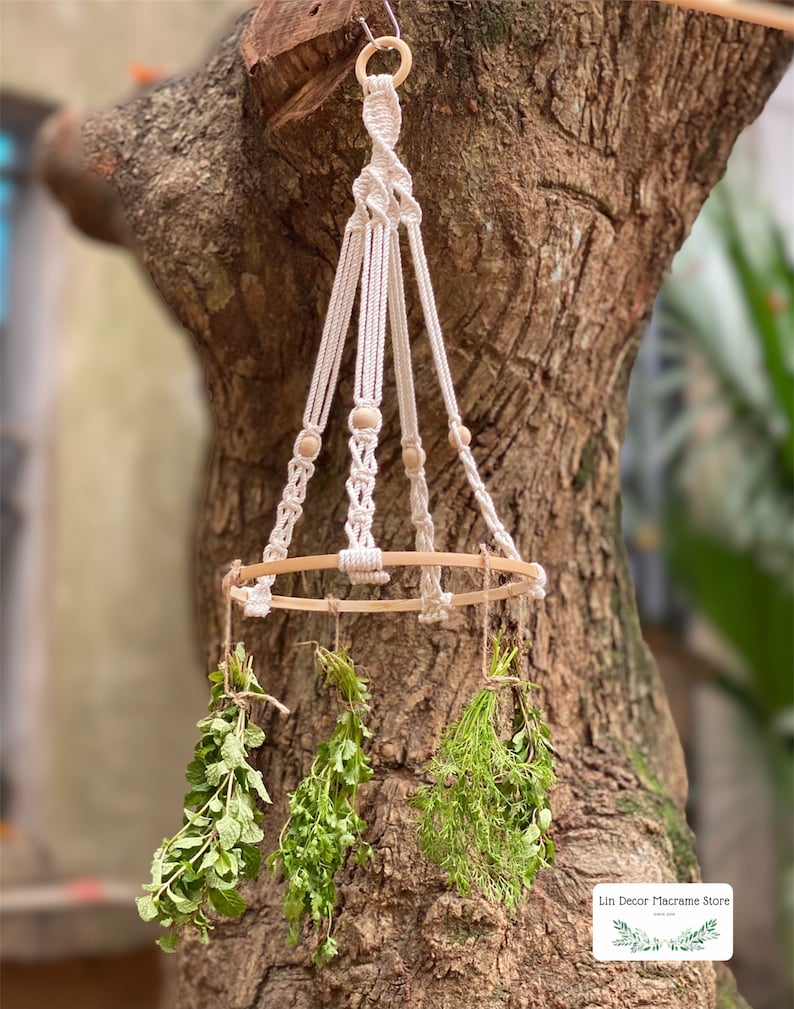 May include: A white macrame hanging planter with a wooden ring and wooden beads. The planter is hanging from a tree branch and has three bundles of dried herbs hanging from it. The text "Lin Decor Macrame Store" is visible in the bottom right corner of the image.
