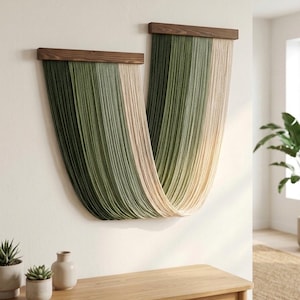 May include: A wall hanging consisting of two horizontal dark brown wooden bars from which long, vertical strands of yarn are suspended. The yarn creates a draped, U-shaped pattern in alternating shades of forest green, sage green, and cream.