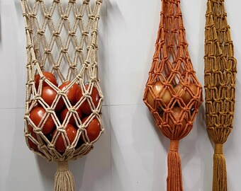 May include: Three macrame hanging fruit baskets in beige, orange, and mustard yellow. The beige basket holds red tomatoes, while the orange and yellow baskets are empty. Each basket has a tassel at the bottom and a wooden ring or rod at the top.