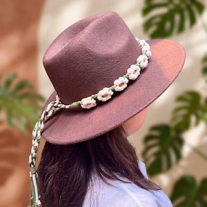 May include: A mauve fedora hat featuring a white daisy chain and a braided strap with a tassel. The hat has a wide brim and a classic crown. The daisy chain is made of white flowers with yellow centres and green leaves.