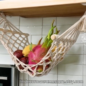 May include: A macram&eacute; fruit hammock, suspended from a wooden shelf, filled with colourful fruits. The hammock is made of off-white woven cotton, with a wooden dowel for support. Fruits include dragon fruit, mangoes, and other produce. The Etsy shop URL is visible at the bottom.