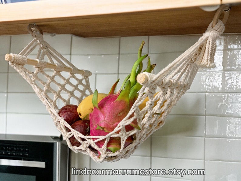 May include: A macram&eacute; fruit hammock, suspended from a wooden shelf, filled with colourful fruits. The hammock is made of off-white woven cotton, with a wooden dowel for support. Fruits include dragon fruit, mangoes, and other produce. The Etsy shop URL is visible at the bottom.