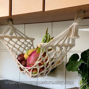 May include: A cream-coloured macrame fruit hammock, suspended from a wooden cabinet, filled with colourful fruit. The hammock is made of woven cotton, with a wooden dowel at the top. The fruit includes dragon fruit, plums, and bananas. The Etsy shop name is visible.