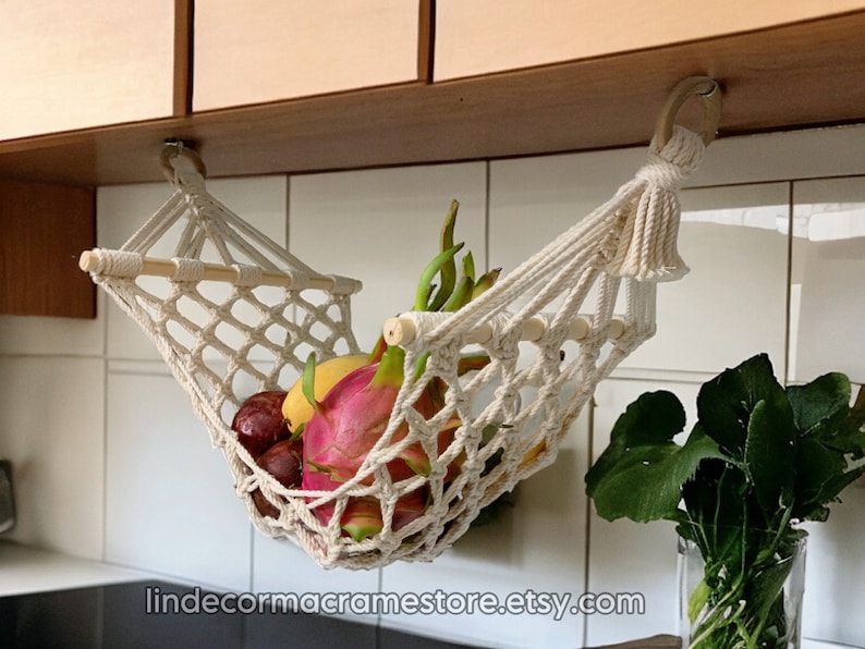 May include: A cream-coloured macrame fruit hammock, suspended from a wooden cabinet, filled with colourful fruit. The hammock is made of woven cotton, with a wooden dowel at the top. The fruit includes dragon fruit, plums, and bananas. The Etsy shop name is visible.