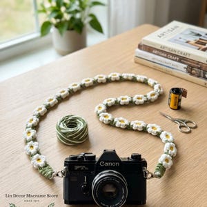 May include: A black Canon FTb camera with a handmade crochet camera strap featuring a repeating pattern of white daisy flowers with yellow centers and green stems. The strap is connected to the camera body with silver metal clips and rings.