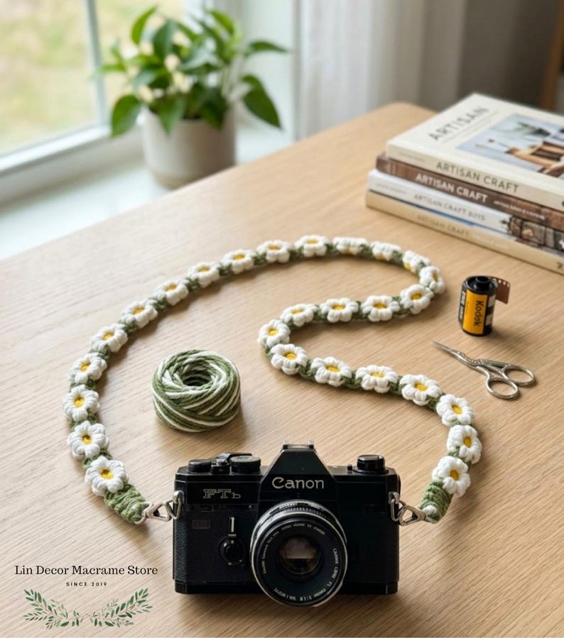 May include: A black Canon FTb camera with a handmade crochet camera strap featuring a repeating pattern of white daisy flowers with yellow centers and green stems. The strap is connected to the camera body with silver metal clips and rings.