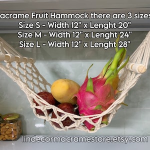 May include: A cream-coloured macrame fruit hammock, with wooden dowels, filled with fresh fruit. The hammock is suspended from the ceiling. The text on the image indicates three sizes: S (30cm x 51cm), M (30cm x 61cm), and L (30cm x 71cm).