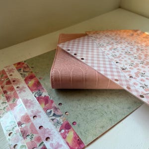 May include: A pink faux-leather planner with a crocodile texture, open to reveal floral and gingham patterned inserts. Several floral-patterned dividers are visible, along with a green floral background. The planner is on a white surface.