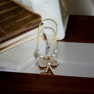 May include: A gold-toned metal paperclip featuring a decorative charm attachment. The charm consists of four white faux pearls and a central gold bead, finished with a dangling bow-shaped pendant encrusted with small clear crystals.
