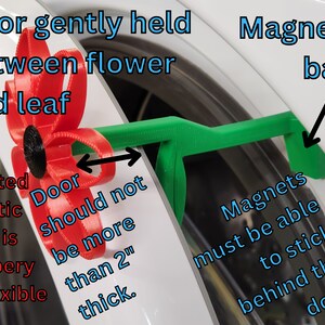 May include: A 3D printed red flower with black center is used to hold a green door stop with a magnetic base. The door stop is designed to hold a door open that is no more than 2 inches thick. The text "Door should not be more than 2" thick." is written on the image.