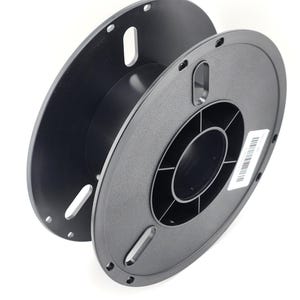 May include: An empty black plastic filament spool featuring a circular design with a central hub, radial support ribs, and elongated slots along the outer rim. A small white rectangular label with a barcode is affixed to the side of the spool.