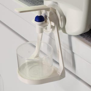 May include: White plastic water dispenser with a blue knob and a clear cup attached. The dispenser is mounted on a white surface.