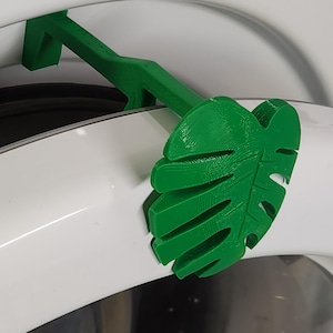 May include: A bright green, 3D-printed leaf-shaped laundry door prop. The prop is designed to hold a washing machine door ajar. The leaf design is a Monstera leaf. The prop is made of plastic and is attached to the washing machine door.