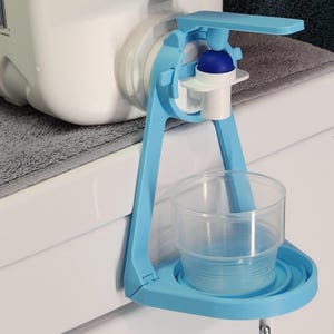 May include: A blue plastic water bottle dispenser with a white spout and a cup holder. The dispenser is attached to a white surface with a gray towel underneath.