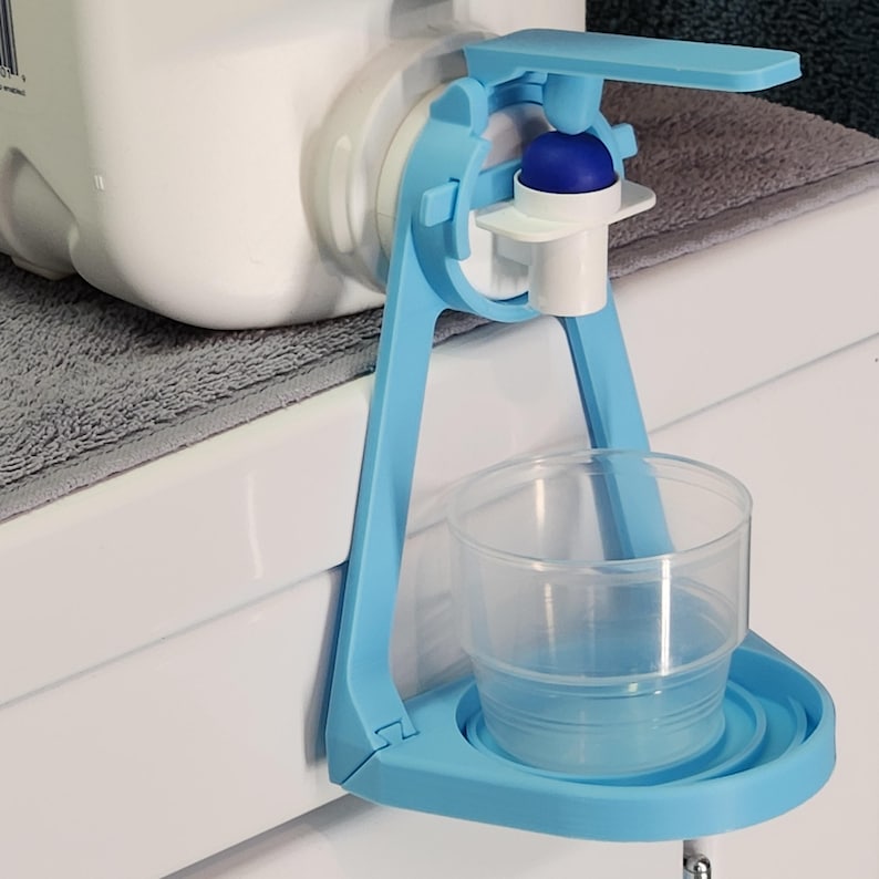 May include: A blue plastic water bottle dispenser with a white spout and a cup holder. The dispenser is attached to a white surface with a gray towel underneath.