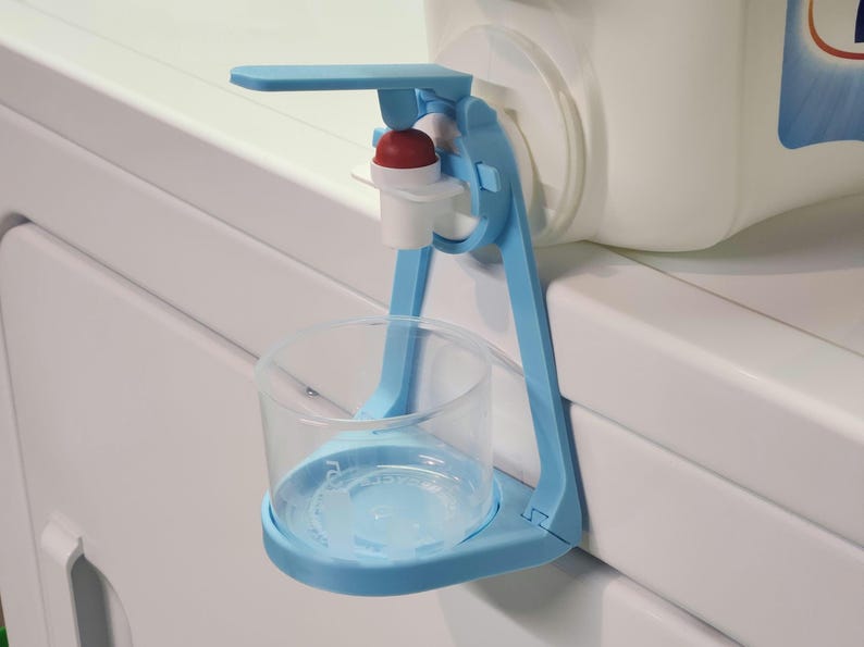 May include: A blue plastic dispenser with a red button and a clear cup holder. The dispenser is attached to a white container.