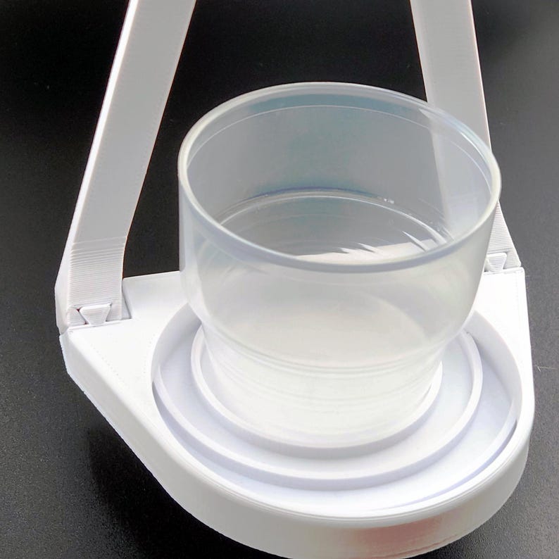 May include: White plastic cup holder with a clear plastic cup inside. The holder has a circular base with a raised lip and a triangular frame.