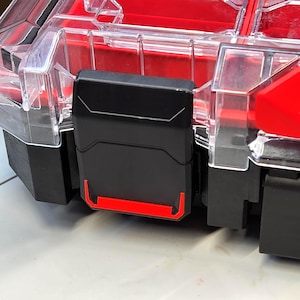 Peut inclure: Gros plan sur une boîte de rangement en plastique noire et rouge. La boîte a un couvercle transparent et un loquet noir avec des accents rouges. L'intérieur de la boîte est rouge.