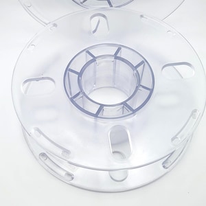 May include: Clear plastic spool with a central hub and multiple circular and oblong openings. The spool is designed to hold filament or wire, and is likely used in 3D printing or similar applications.