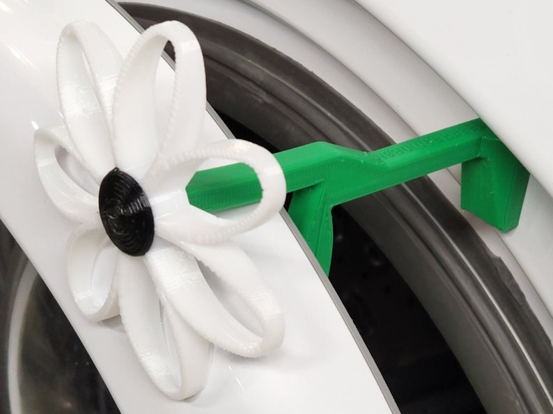 May include: A 3D printed white flower with a black center attached to a green plastic hook. The hook is designed to be used as a tool for opening a washing machine door.