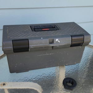 May include: A gray toolbox with black latches and a red button on the handle. The top has a textured pattern. A black rubber ring sits in front of the toolbox.