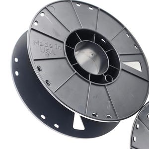 May include: Close-up of a black plastic spool with the text "Made in USA" visible. The spool has a circular design with multiple holes around the edge and is likely used for storing filament or wire.